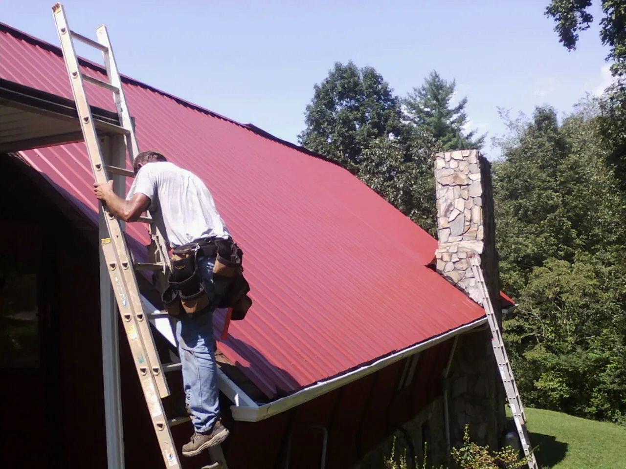Roofer on a ladder beside a red metal roof and stone chimney — Soffit & Fascia Repair by Roofing Brothers in Oakhurst
